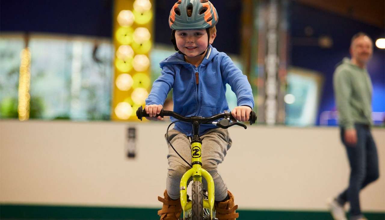 Child on a small yellow bicycle rides forward, smiling, while wearing a helmet; indoor venue with soft lighting and a blurred adult behind. Text: 2 on the bike’s front.