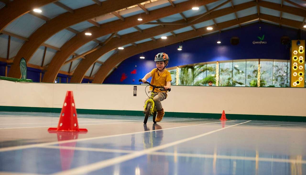 Child on a small balance bike rides between red cones, wearing a helmet, on an indoor court under an arched wooden roof; blue wall, windows, climbing feature. Text: “Center Parcs”, “E11”.