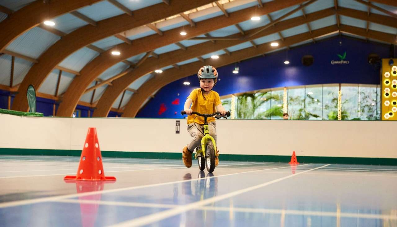 Child on a small bike rides between orange cones, wearing a helmet, on a glossy indoor court beneath arched beams; greenery visible behind glass. Visible text: 2, Center Parcs.