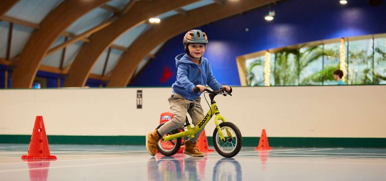 Child on a small green balance bike rides between orange cones on an indoor court under arched wooden beams, wearing a helmet and boots. Text: woom; 11.
