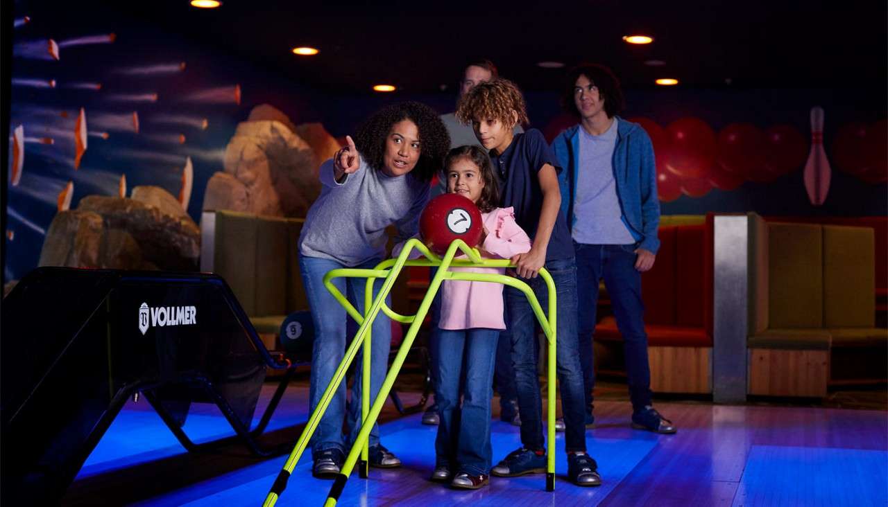 Child prepares to roll a red bowling ball using a neon-green ramp, guided by adults, in a dimly lit bowling alley. Text: VOLLMER; 9.