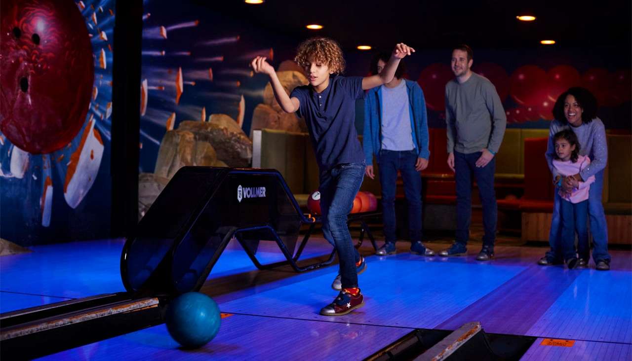 Boy bowls a teal ball down a lane, stepping forward; family watches behind at a neon-lit bowling alley with mural of a red ball striking pins. Text: VOLLMER.