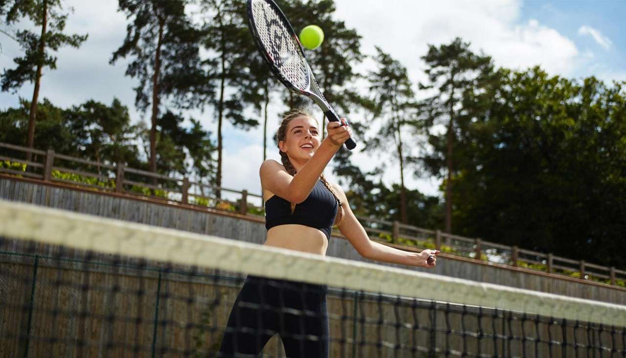 Tennis player hits a forehand volley, smiling as the ball approaches the racket, on an outdoor court with the net in foreground and tall trees against a bright sky.