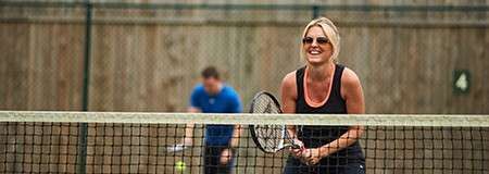 Woman tennis player smiles while gripping a racket at the net; background player readies a ball. Scene: outdoor court with chain-link fence, wooden barrier, and a court sign reading "4".