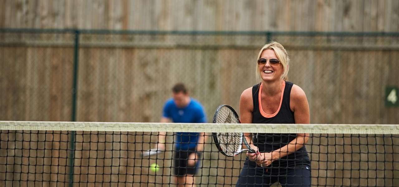 Woman in sunglasses holds a tennis racquet ready at the net, smiling; partner in blue behind prepares a shot; outdoor court with chain-link fence, wooden backdrop, and sign reading "4".