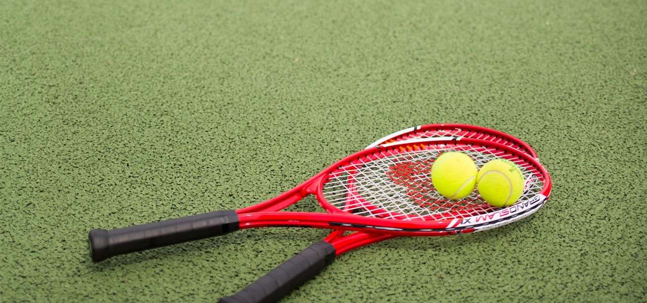 Two red tennis rackets lie crossed, holding two yellow tennis balls, on a green textured court surface. Text on rim: GRAND SLAM X1.