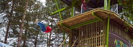 A person zip-lines outward, legs up, from a high wooden adventure tower; an attendant waves from the platform; tall pine trees surround the structure. Sign reads ACT.
