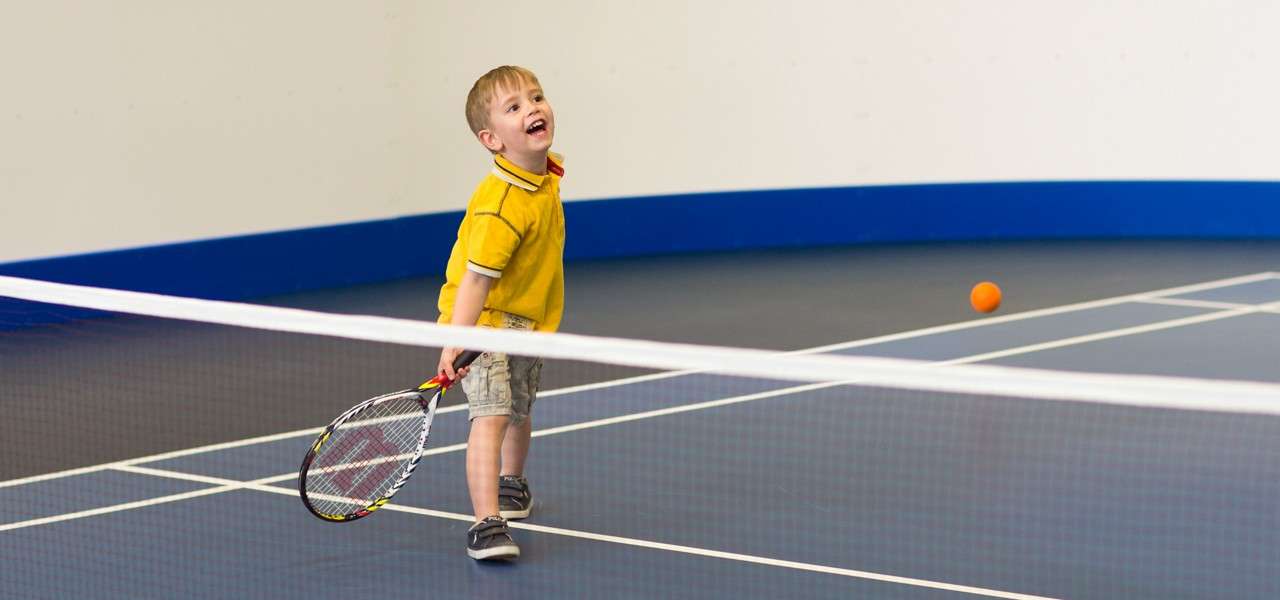 Young child holds a tennis racket and smiles after a swing, watching an orange ball bounce across a net on an indoor court with blue border lines.