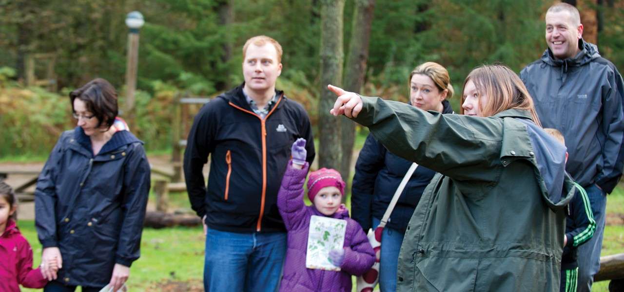 Guide points and speaks; adults and children listen, bundled in jackets; forest clearing context with trees and wooden benches visible in the background on a cool day.