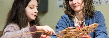 A child and an adult weave thin wooden reeds, hands moving through a lattice, while sitting together at a table in a well-lit indoor room.