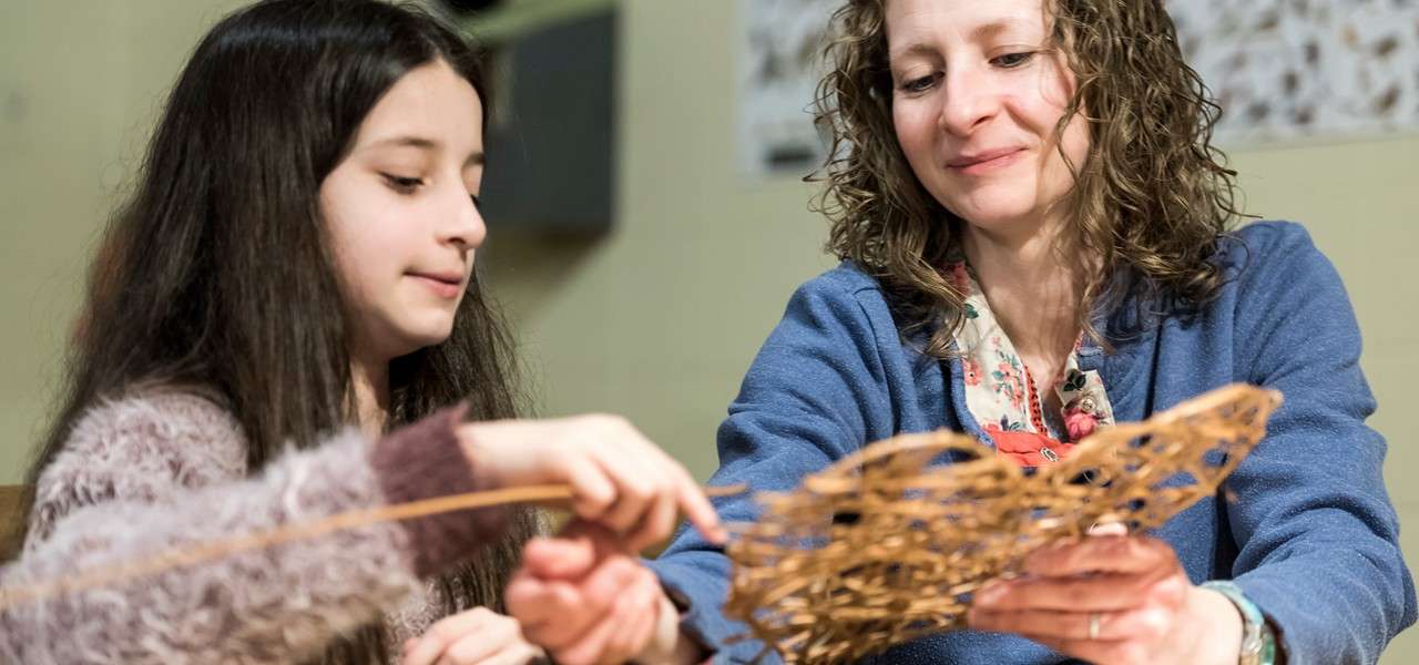 Woven craft—two people weave thin wooden strips, shaping a lattice with their hands at a table in a well-lit indoor workspace.