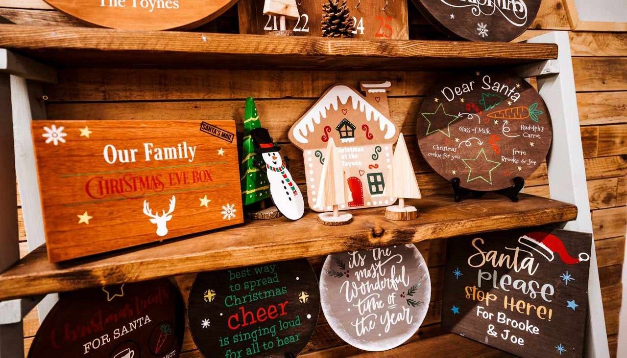 Wooden Christmas signs—arranged on rustic shelves—decorate a cozy shop display.

Text: The Toynes; 21 22 24 25; Our Family; CHRISTMAS EVE BOX; SANTA’S MAIL; Christmas at the Toynes; Dear Santa; Glass of Milk; Rudolph’s Carrot; Christmas cookie for you; from Brooke & Joe; best way to spread Christmas cheer is singing loud for all to hear; it’s the most wonderful time of the year; Santa please STOP HERE! For Brooke & Joe.