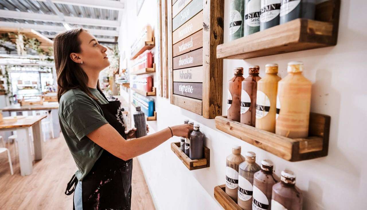 Woman selects a stain bottle from wall shelves in a bright craft studio; color board reads: sage, rosewood, redwood, chocolate, khaki, expresso; bottle labels: STAIN.