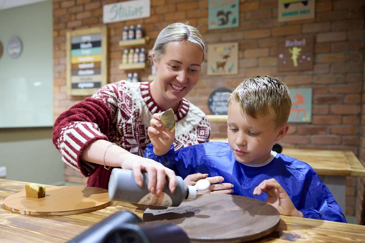 Woman guides child pouring paint onto a wooden round; child holds a sponge, both working at a table in a craft workshop with brick wall, shelves, and signs. Text: “Stains”, “Rebel”.