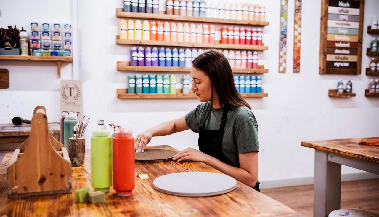 Woman artist sands a round wooden board with a sponge, seated at a worktable; surrounding shelves display rows of colorful paint bottles, brushes, and craft supplies in a bright studio.