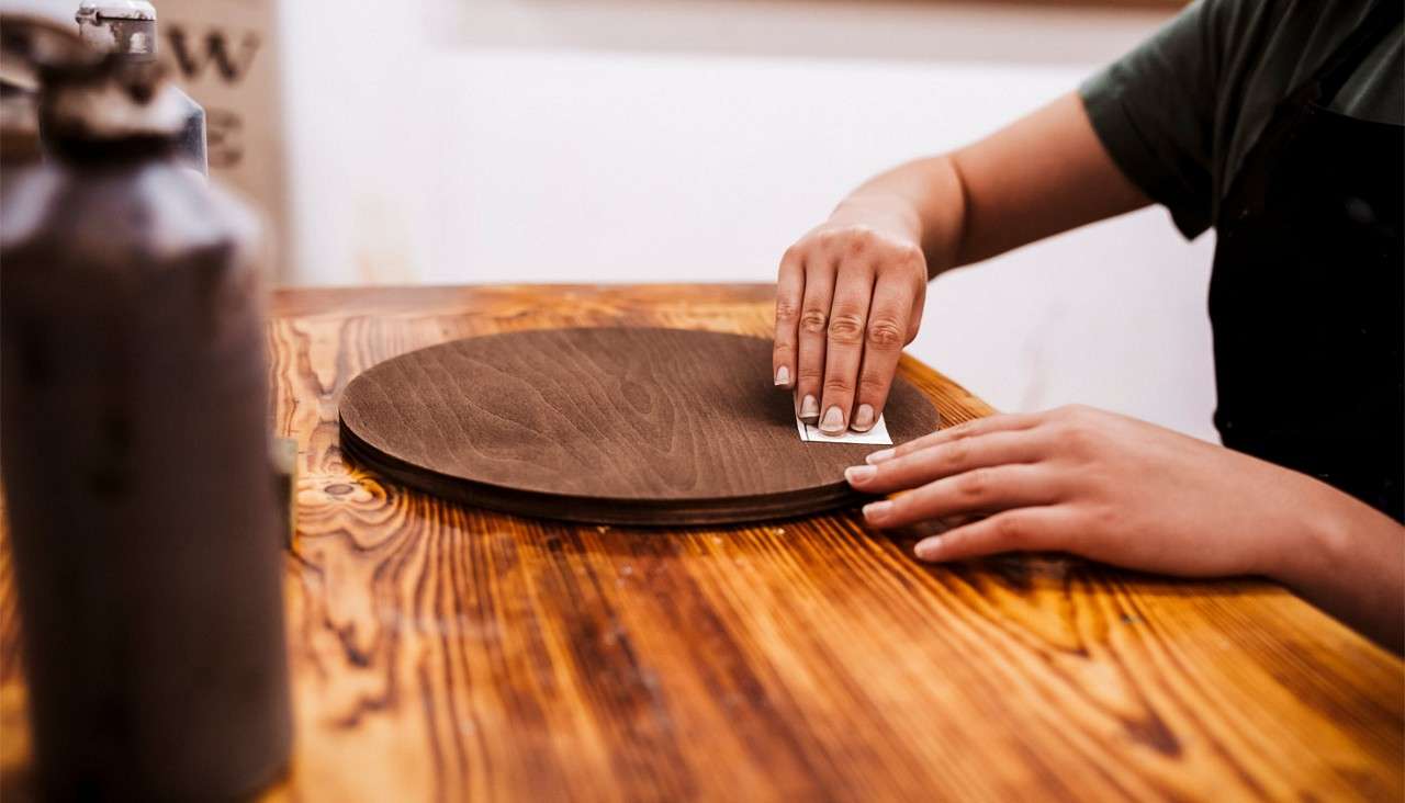 Round wooden board being sanded by two hands with a small sanding block, resting on a rustic workbench in a workshop, with blurred containers nearby and a person wearing a dark apron.