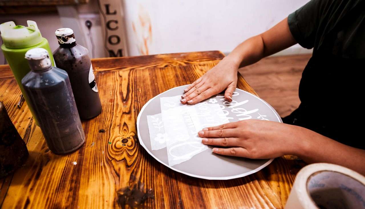 Hands smooth a stencil onto a round gray sign, applying transfer paper; on a wooden worktable with paint bottles and tape. Visible text: LCOM, tesa.