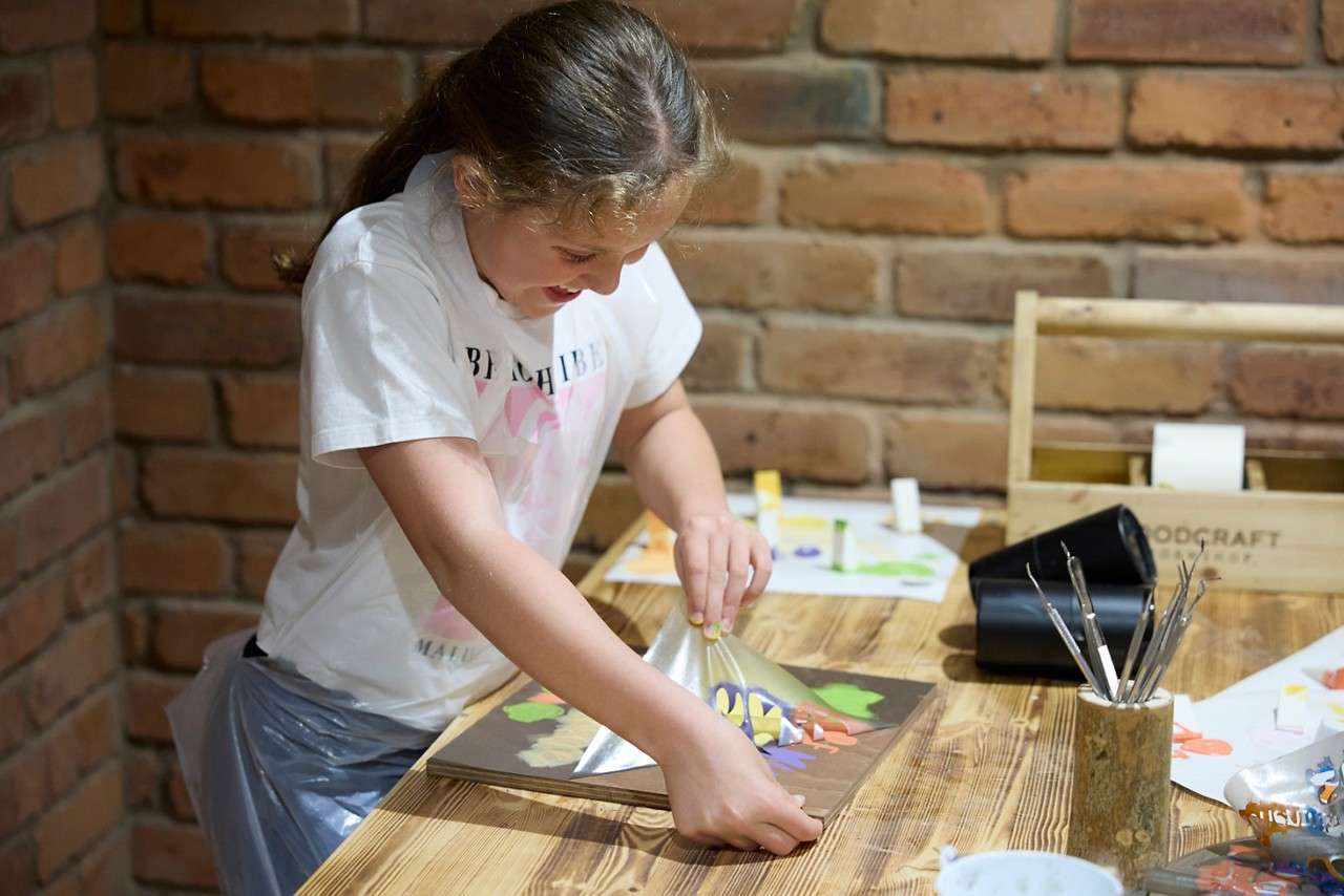 Girl peels a shiny stencil from wet paint on a board, working at a wooden table in a brick-walled studio with brushes and tools. Text: WOODCRAFT; shirt: …HIBI… (partially obscured).
