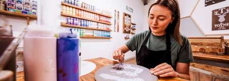 Person dots paint onto a round ceramic plate using a small tool, at a wooden table in a craft studio with shelves of paint bottles and wall décor in the background.