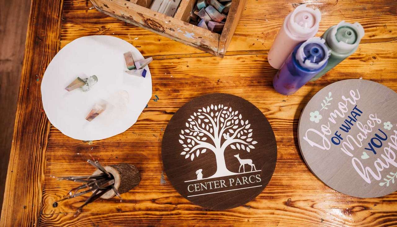 Wooden craft plaques sit on a workshop table, surrounded by brushes, pastels, and paint bottles. Text: CENTER PARCS. Do more OF WHAT Makes YOU happ.