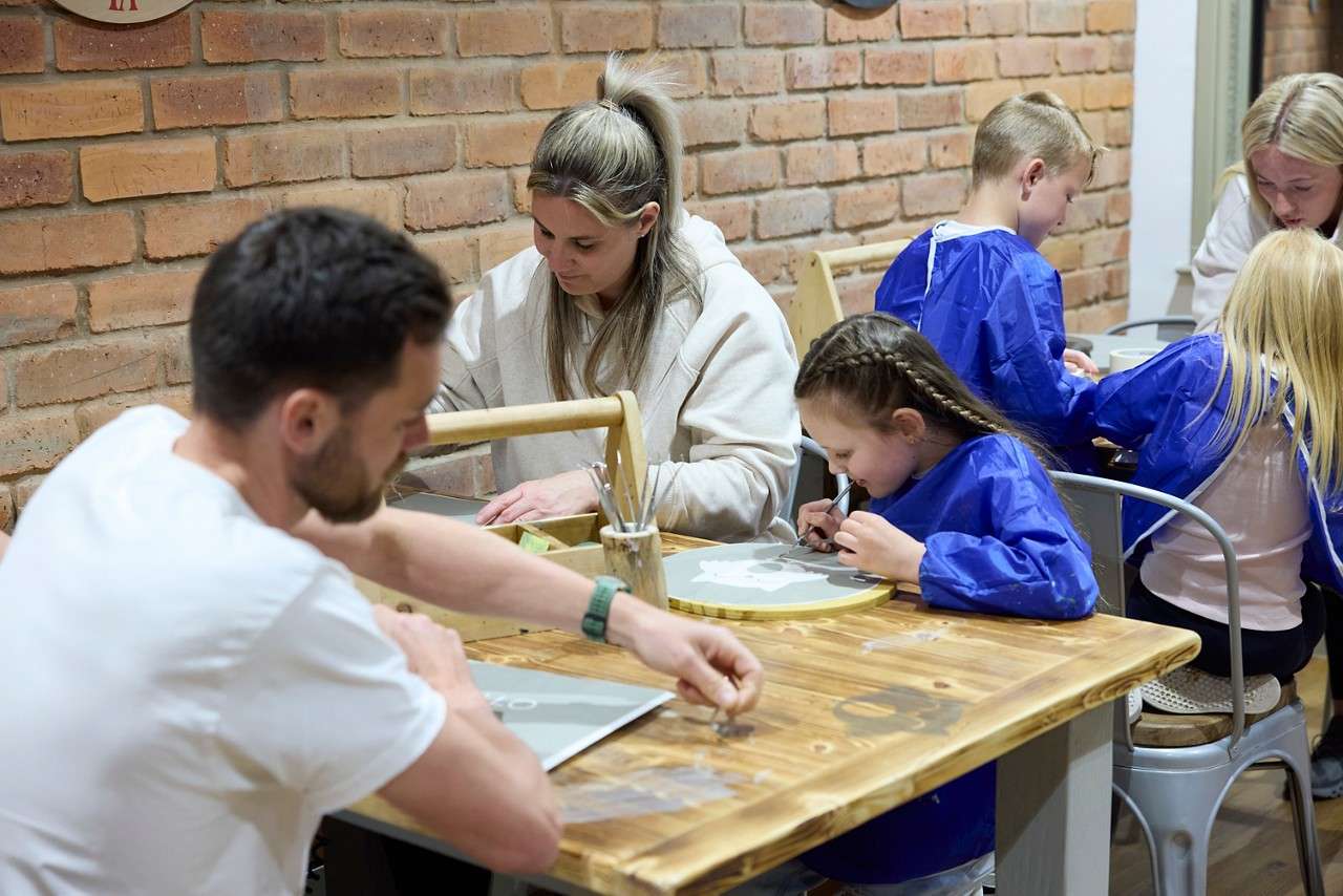 Children and adults paint crafts at a wooden table, concentrating on designs using brushes and palettes. Smock-wearing kids work beside supervising adults in a cozy brick-walled art studio.