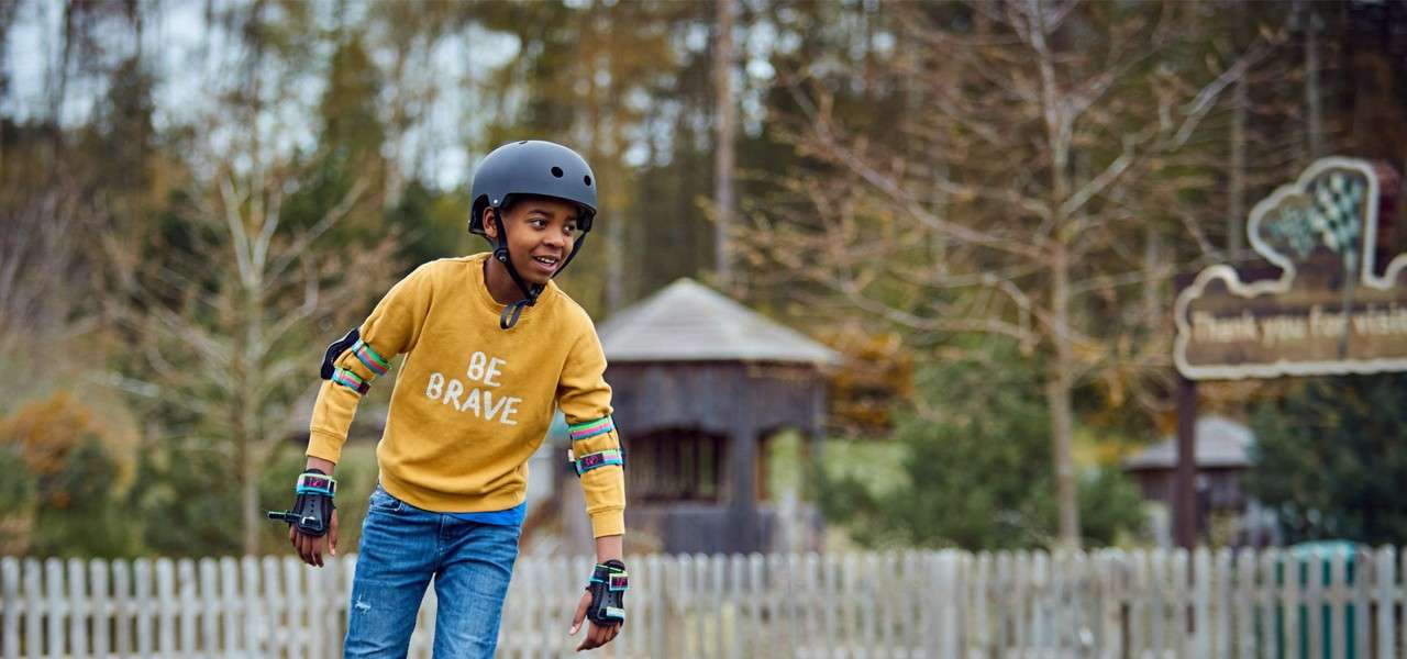 Child in helmet and pads moves forward smiling, outdoors near a white picket fence and trees; sweatshirt reads BE BRAVE; background sign reads Thank you for visiting.