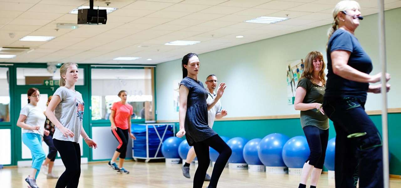 Group of adults perform dance steps, following a headset-wearing instructor, in a bright fitness studio with wooden floor, teal walls, ceiling lights, and blue exercise balls lined along the wall.