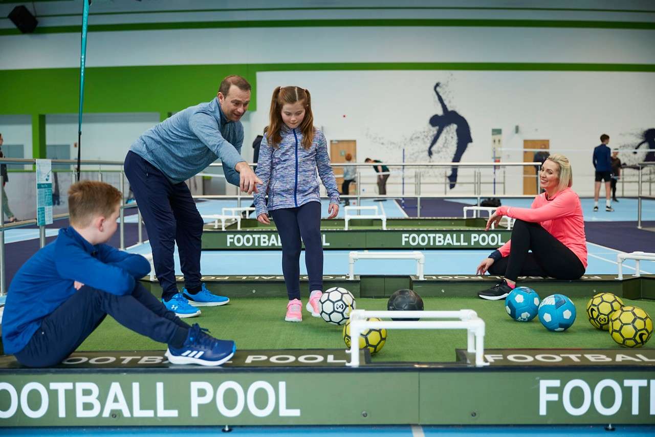 A family competing in football pool. A little girl is about to kick while dad looks over.