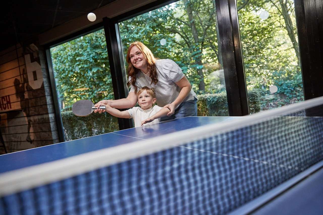 A toddler playing table tennis while mum helps hold the racket