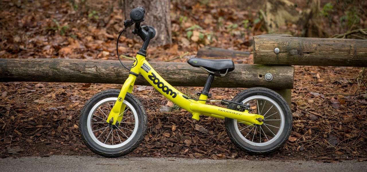 A small yellow balance bike leans against a wooden log fence on a leaf-strewn park path. Visible text: “zooom,” “adventure,” “Y02.”