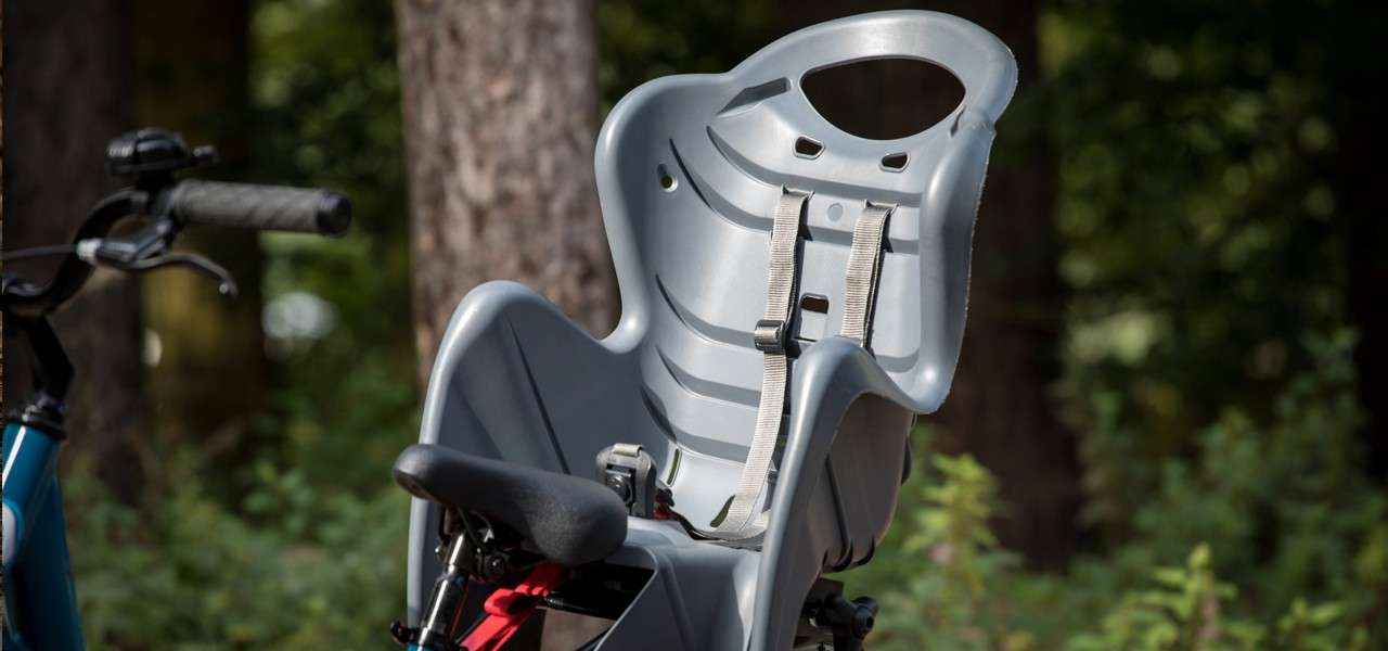 A gray child bicycle seat with shoulder harnesses is mounted behind the saddle, facing forward, while the bike rests in a forested area with trees and foliage in soft focus.