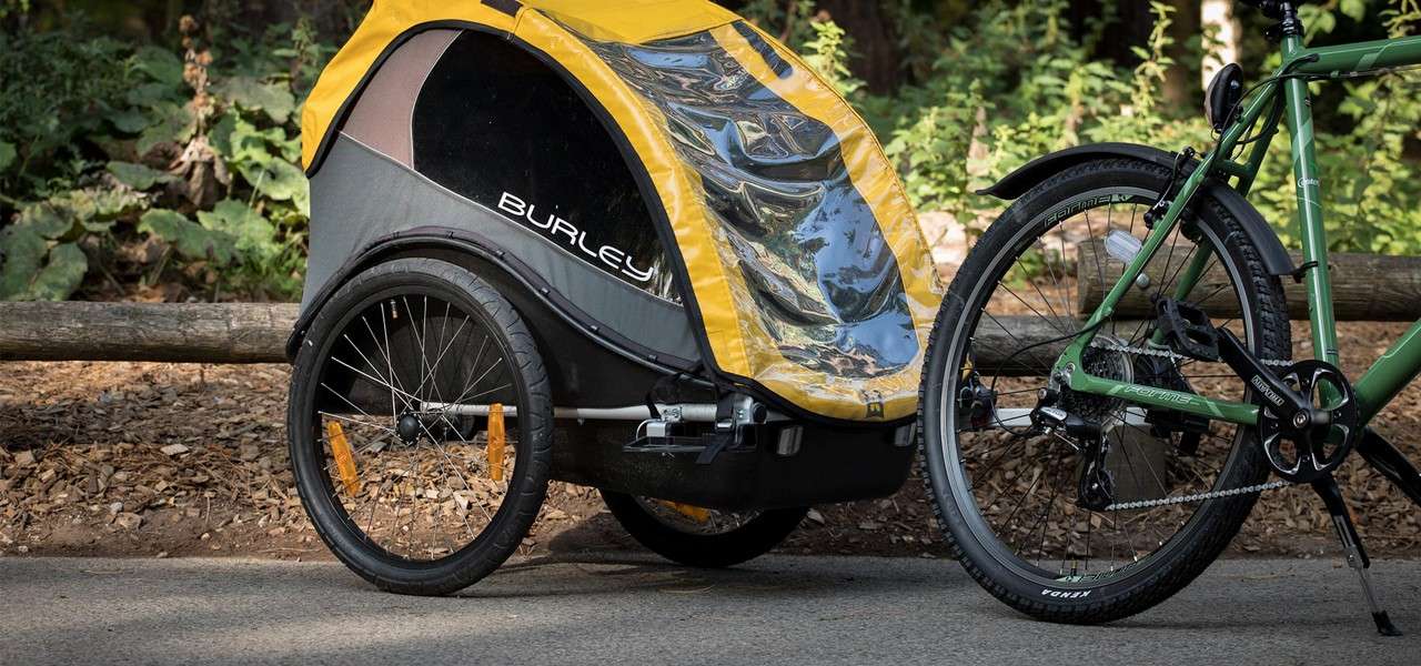 Child bike trailer sits attached to a green bicycle on a paved forest path beside a wooden fence. Text visible: BURLEY on the trailer; KENDA on the tire.