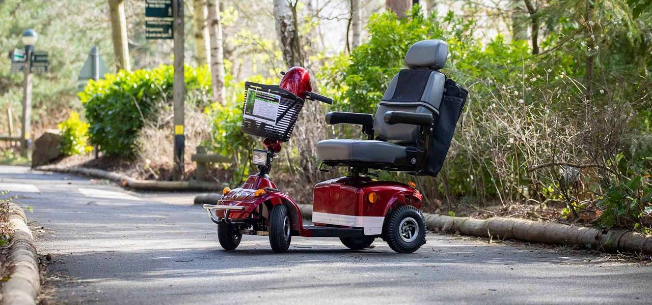 Red mobility scooter sits parked, front basket facing left, on a paved path in a leafy park; trees, shrubs, and wooden edging line the sunlit walkway.