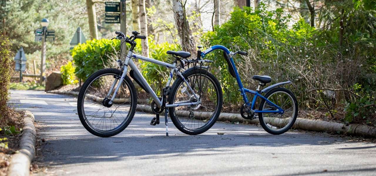 Tandem-style adult bike with child trailer attachment stands upright, linked together, on a shaded paved path bordered by shrubs and trees in a park-like setting.