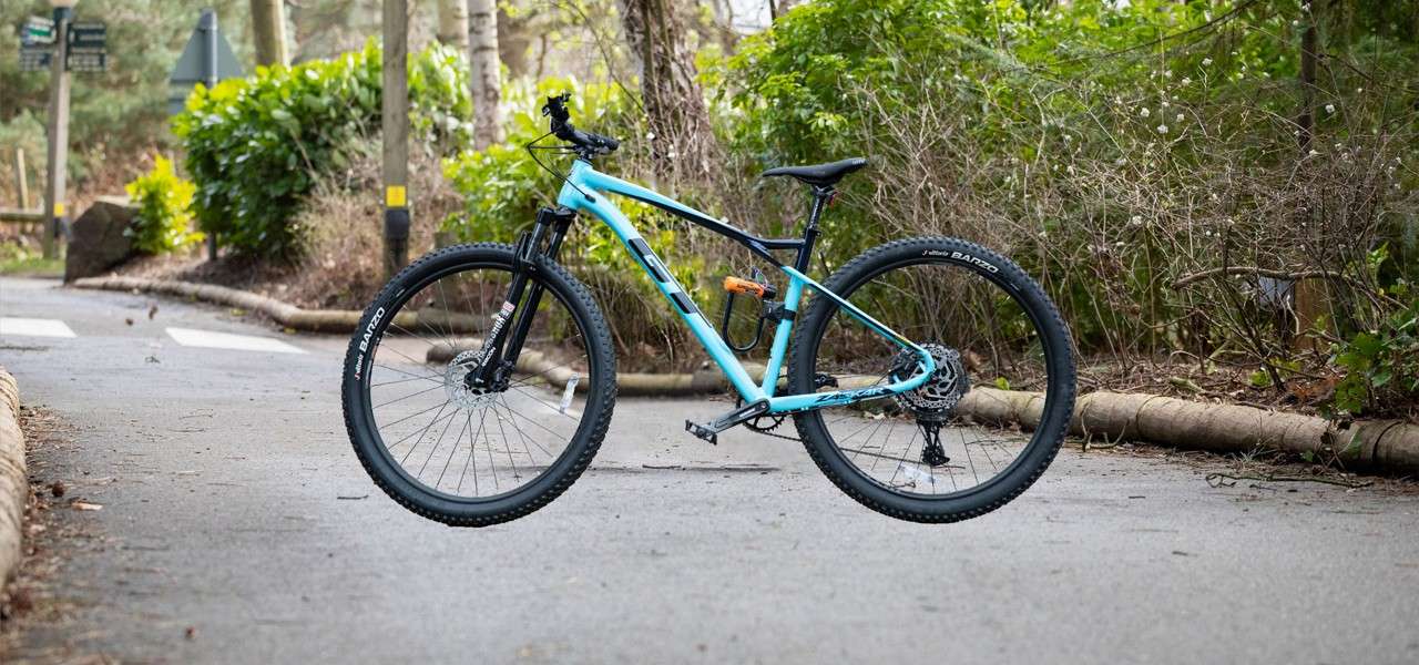 Blue hardtail mountain bike hovering upright on a paved park path lined with greenery and trees. Visible text: GT; ZASKAR; Vittoria BARZO; ROCKSHOX.