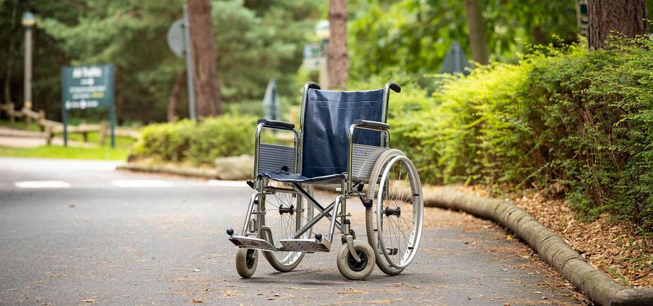 Wheelchair stands empty on a paved path, slightly angled; surrounded by green shrubs and tall trees in a park, with blurred signboards and soft daylight.
