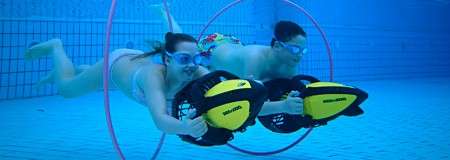 Two swimmers ride yellow underwater scooters, gliding side by side through a pink hoop in a clear blue tiled swimming pool, wearing goggles and swimsuits.