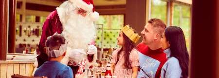 Santa Claus leans toward children, presenting a gift, while a family wearing paper crowns sits at a festive restaurant table, watching and smiling amid holiday decorations and daylight windows.