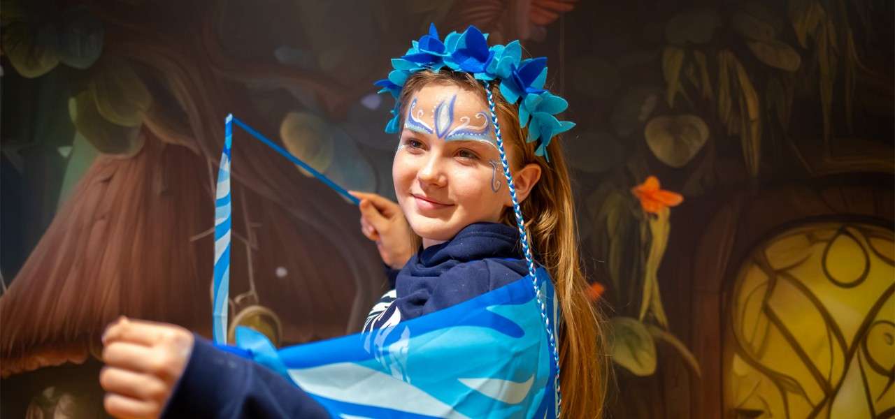 Child with blue floral crown and face paint twirls a blue ribbon, smiling, amid a fantasy forest mural with oversized tree trunk, hanging leaves, a glowing rounded door, and orange flowers.