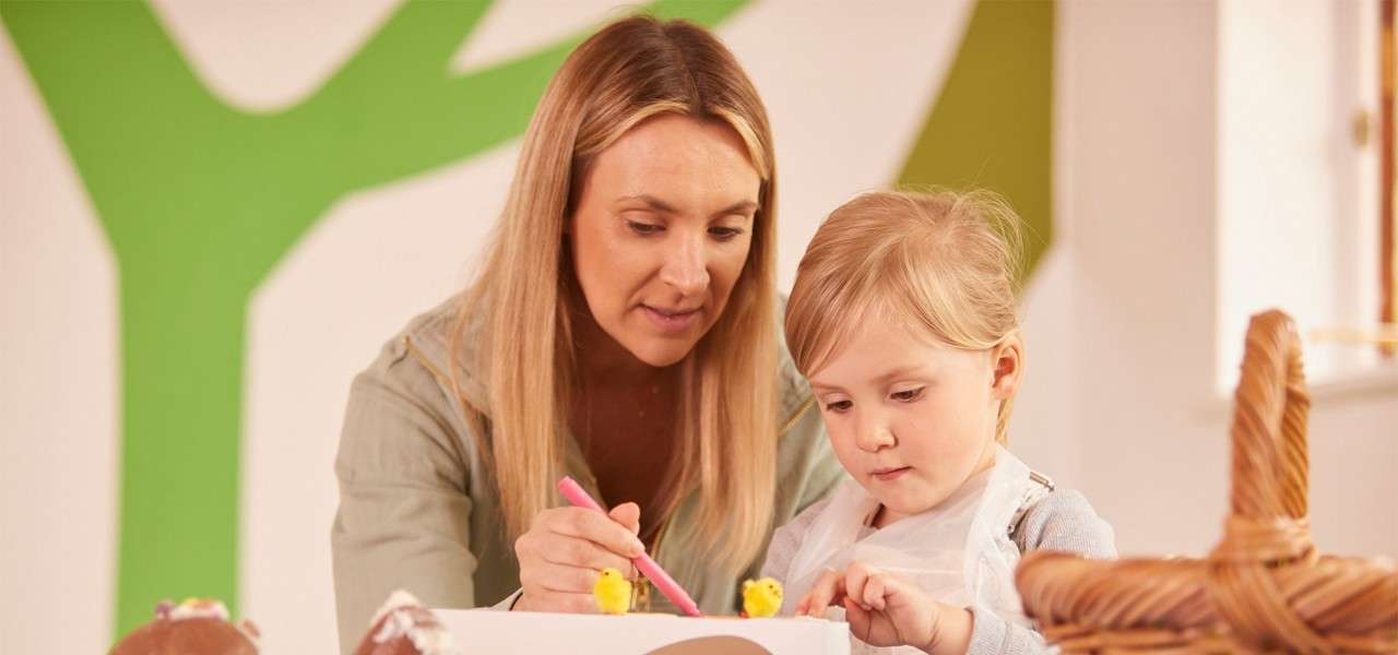 Two people—an adult and a child—decorate a white box, drawing with a pink marker and arranging small yellow chicks, in a classroom with a green tree mural and baskets.