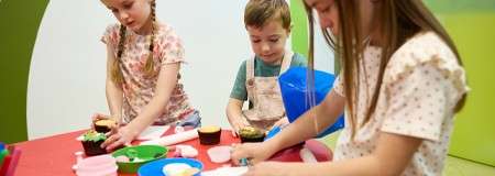 Three children mold colorful playdough, rolling and shaping pieces on a red table; bowls, cutters, and toy cupcakes surround them in a bright classroom with green-and-white walls and soft lighting.
