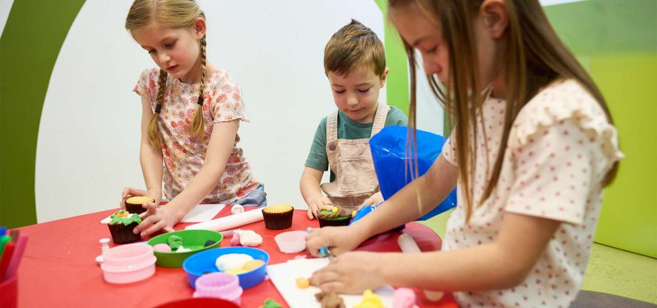 Children decorate cupcakes, rolling and shaping colorful fondant with cutters and bowls, at a red table in a bright classroom play area.