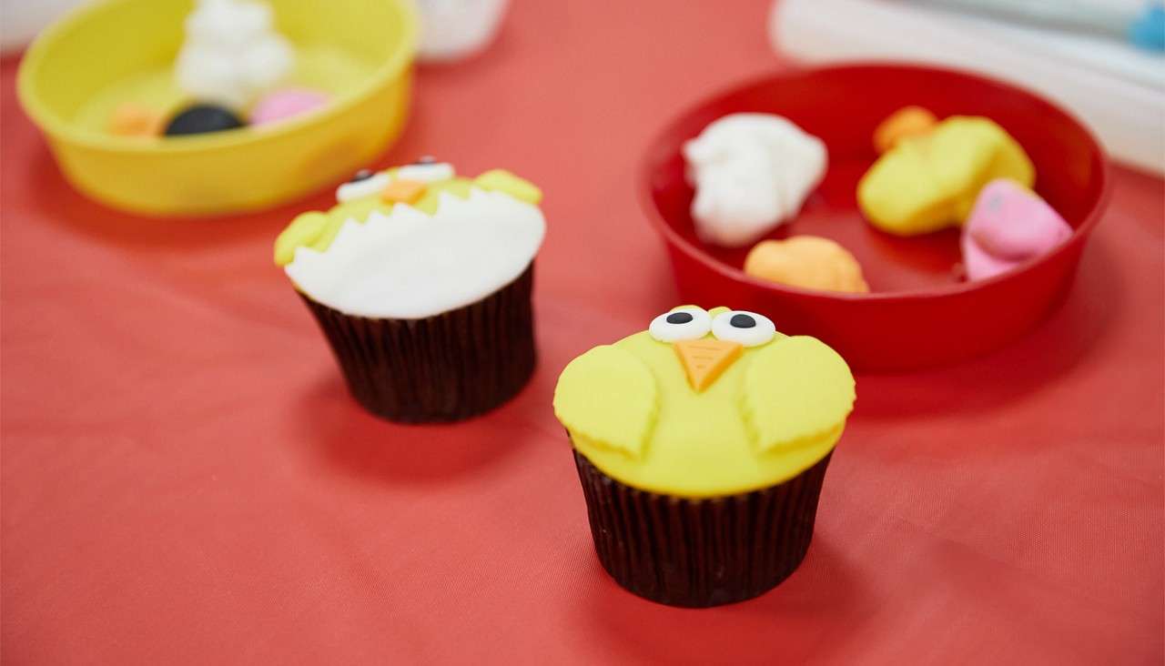 Chick-decorated cupcakes rest on a red tablecloth, while nearby bowls hold colored fondant pieces, indicating a cupcake-decorating activity.
