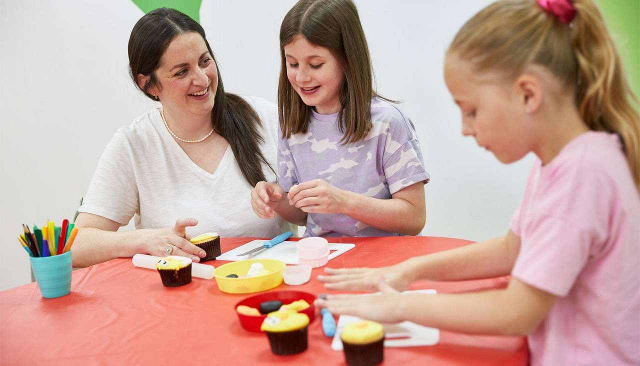 Children and an adult decorate cupcakes, shaping colorful fondant on mats and tools, seated at a red table in a bright classroom, with markers in a cup and cupcakes nearby.