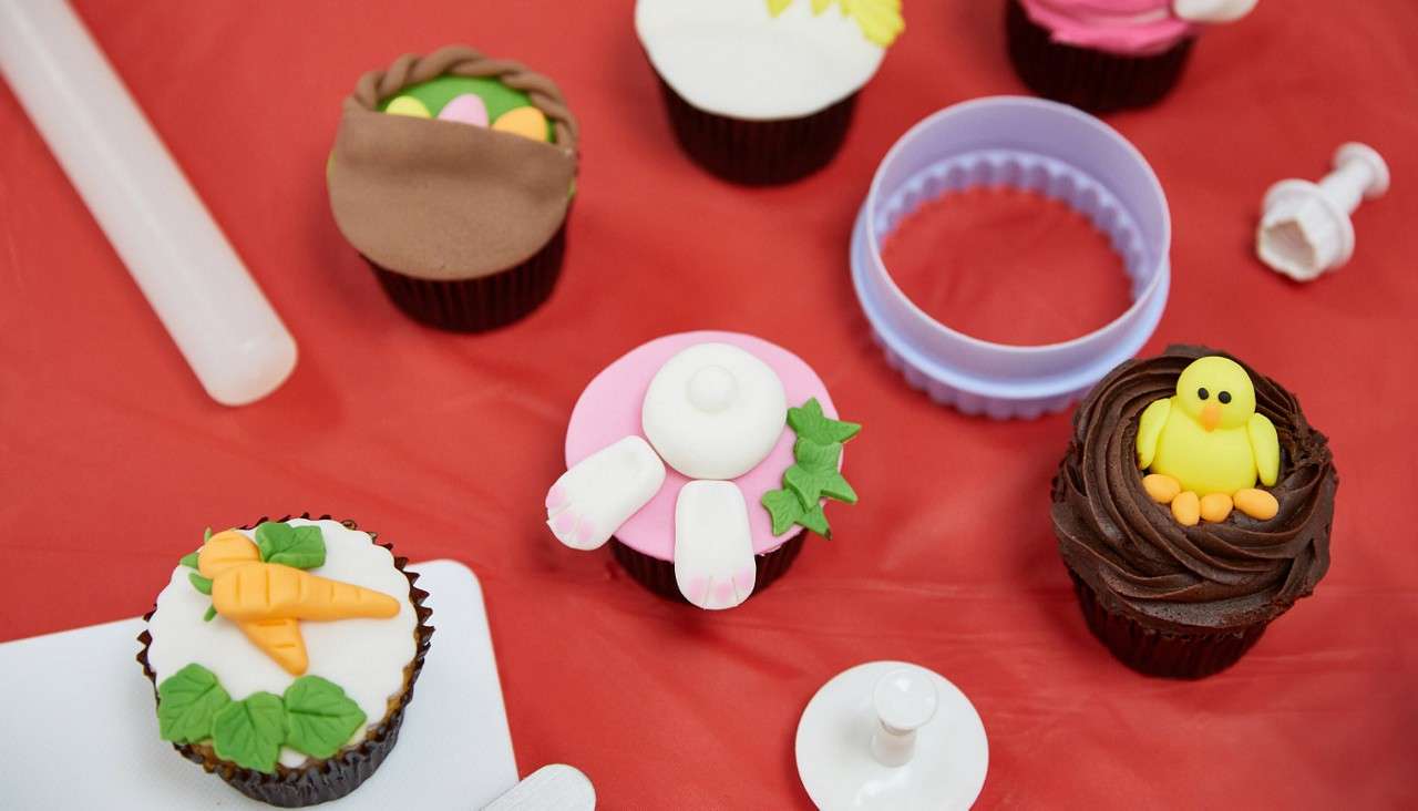Cupcakes display colorful fondant decorations—carrots, a rabbit figure, and a chick—resting on a red tablecloth alongside baking tools, including a rolling pin, cutter rings, and plunger stamps.