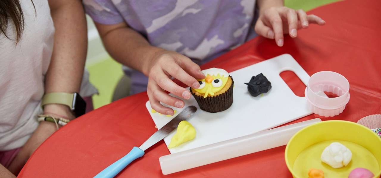 Hands decorate a cupcake with yellow icing and candy eyes on a white cutting board, surrounded by a knife, cookie cutter, and rolling pin on a red table in a craft setting.
