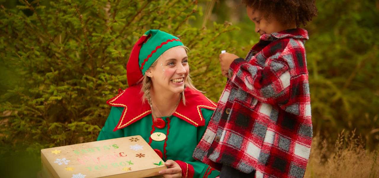 Elf-costumed adult kneels, smiling while holding a wooden box labeled “SANTA’S MAGIC TOY BOX”; child in a red plaid coat examines a small item; scene set among green conifer trees outdoors.