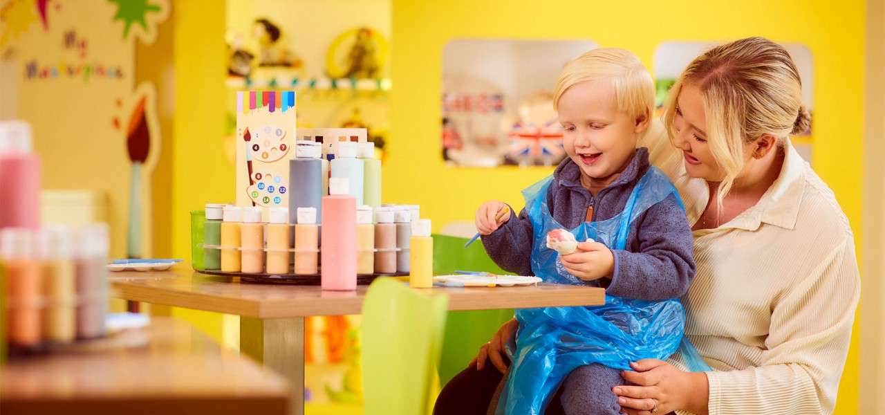Child paints a small ceramic figure, smiling in a blue apron as a woman supports him. A bright craft studio surrounds them with paint bottles, brushes, and colorful decorations.