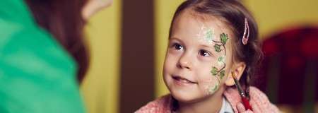 Child with floral face paint smiles and looks up at an adult, sitting indoors in warm light; pink sweater and hair clip visible, green-painted leaves and vines across cheek.