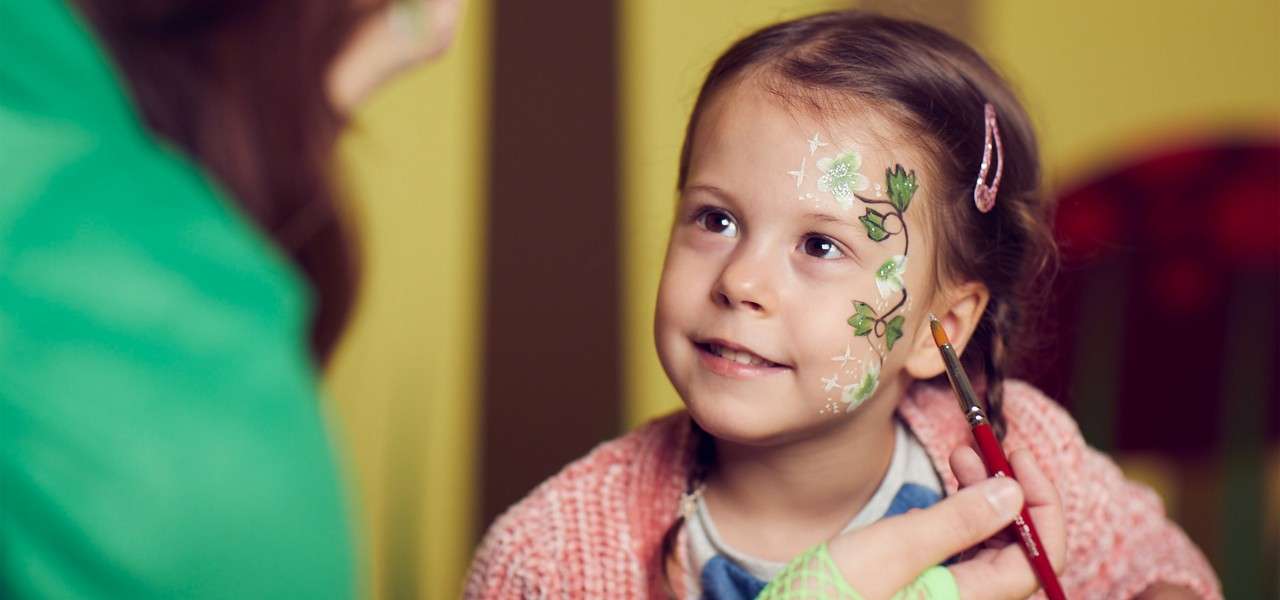 Child receives face painting—artist brushes green leaf and vine design on her cheek—inside a colorful indoor setting; child gazes upward, smiling gently.
